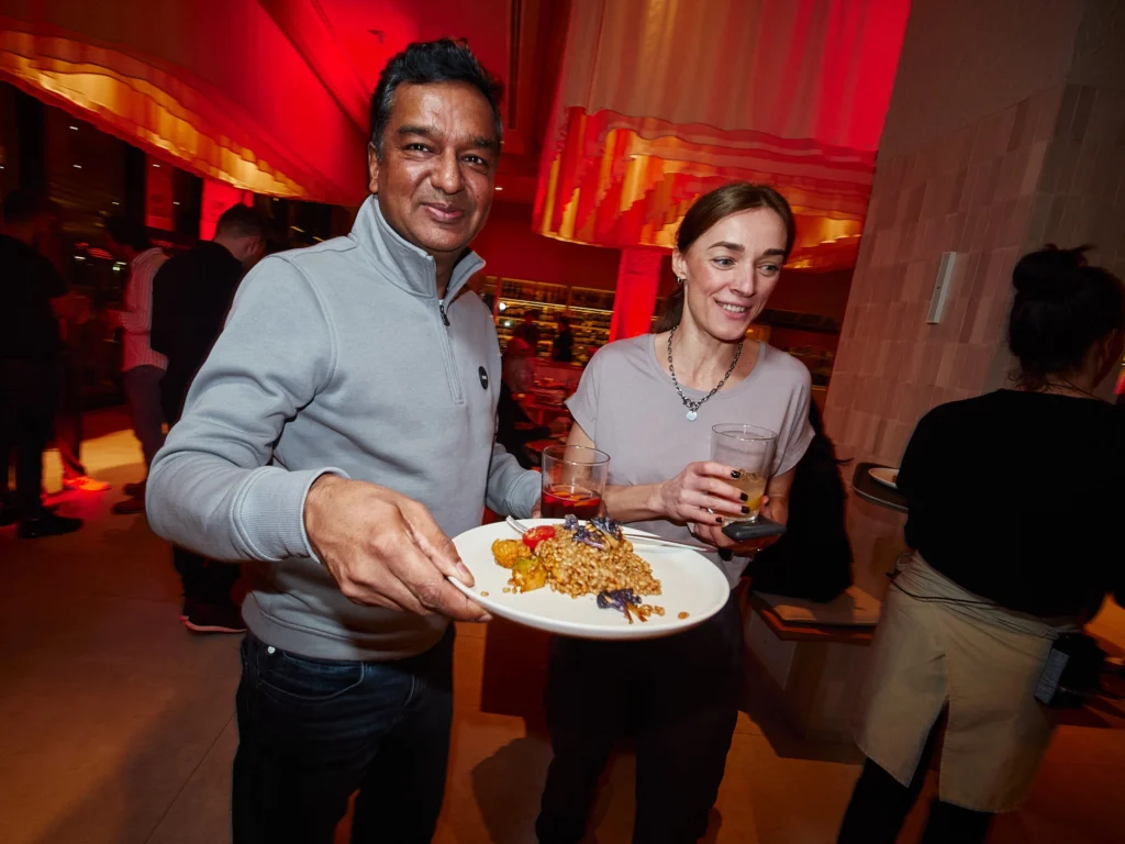 A tech executive looks confidently at the camera holding a plate of Paella during the Odys corporate client evening, with Nuara's sophisticated mood lighting in the background.