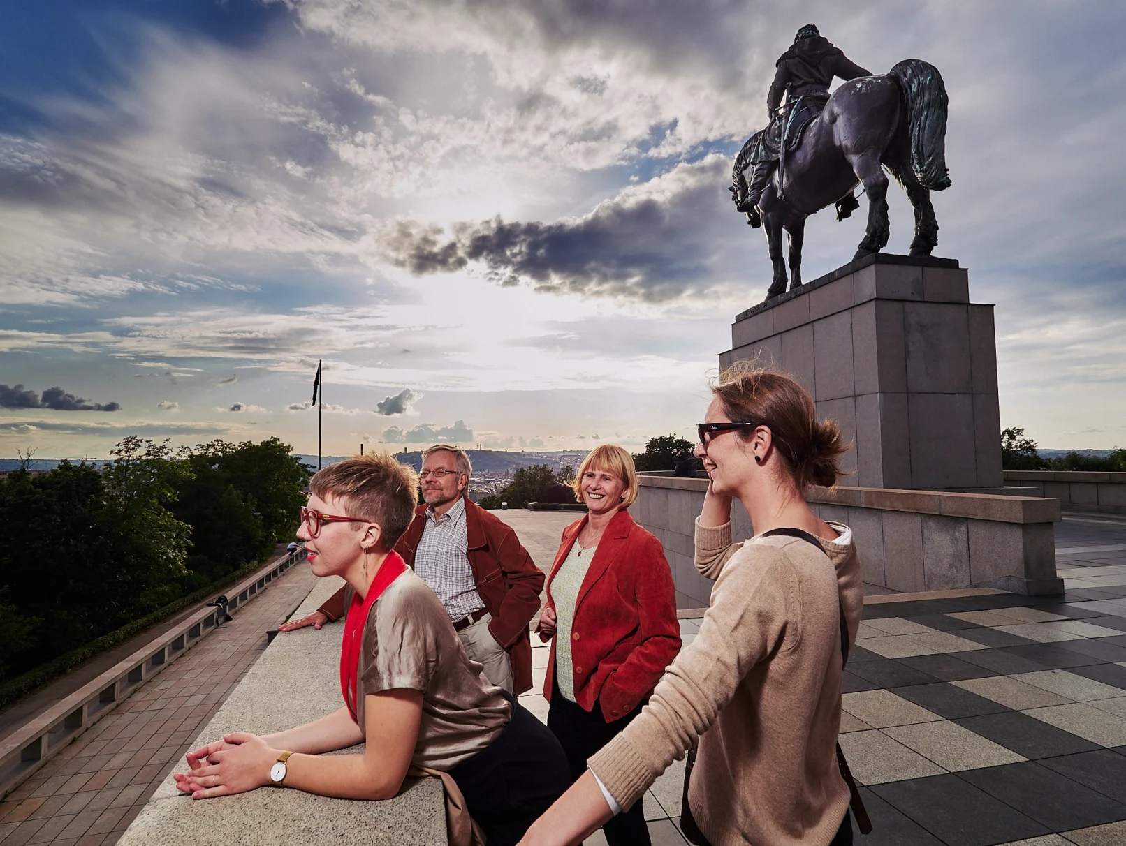 Four travelers stand at a scenic viewpoint in Prague near the Equestrian Statue of Jan Žižka on Vítkov Hill, overlooking the city skyline at golden hour, with dramatic clouds and soft sunlight in the background.