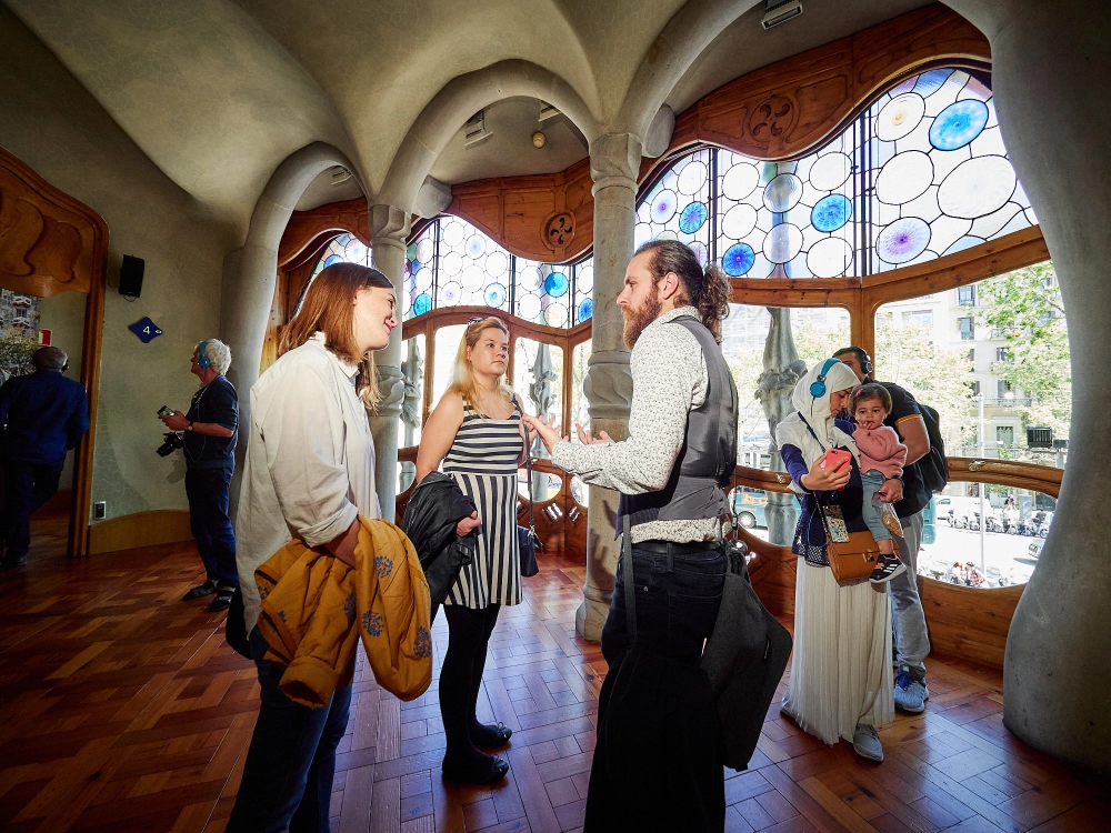 A guide talks to a small group of visitors in a large, ornate room with high ceilings and large windows, characteristic of a modernist palace.